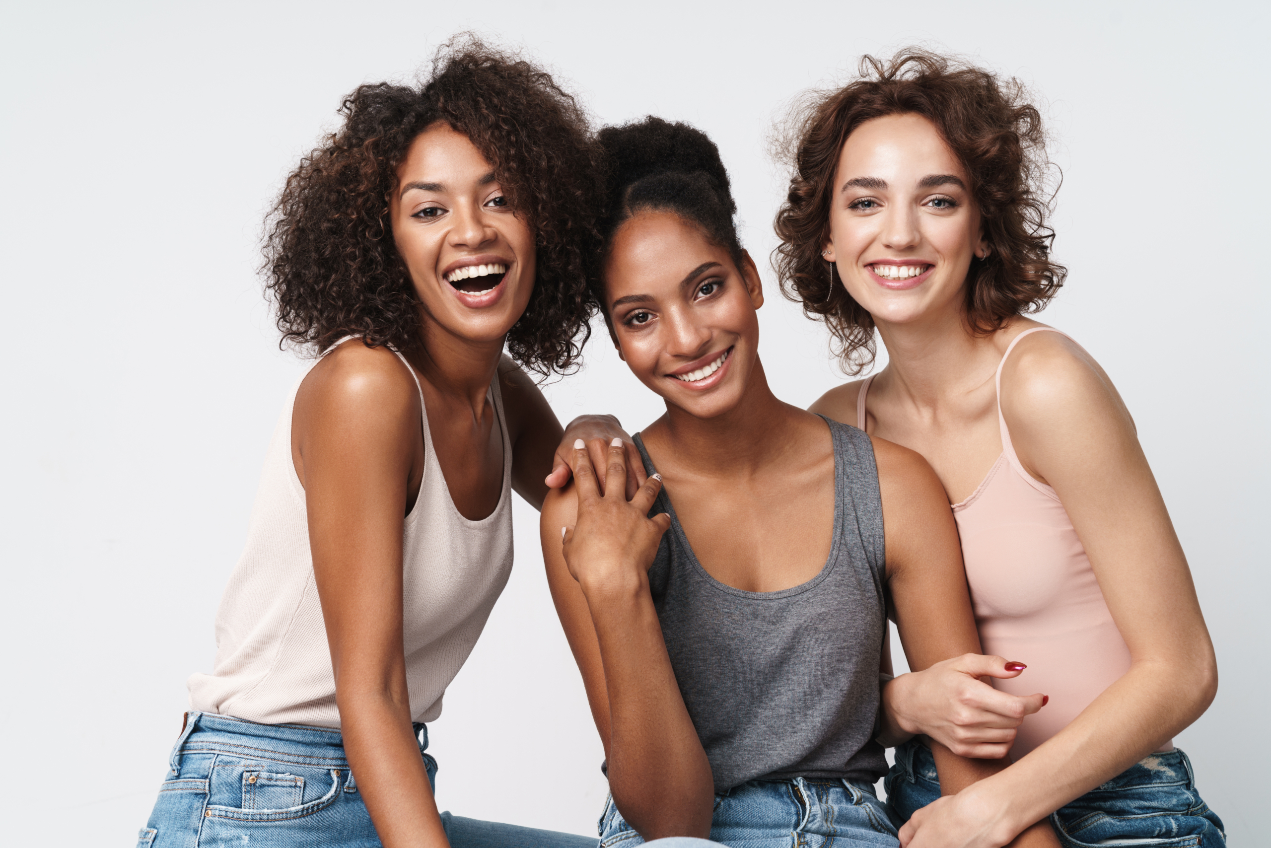 Portrait of three gorgeous multiracial women standing together and smiling at camera isolated over white background