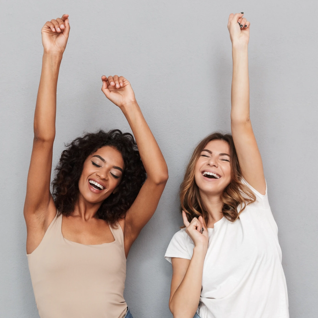 Two women share an exuberant moment of joy as they dance, laughing with their arms raised in the air. The woman on the left has dark curly hair and a tan top, while the woman on the right has straight blonde hair and is wearing a white T-shirt. Their faces are filled with pure happiness and carefree energy, set against a minimalist grey backdrop, emphasising their celebratory mood.