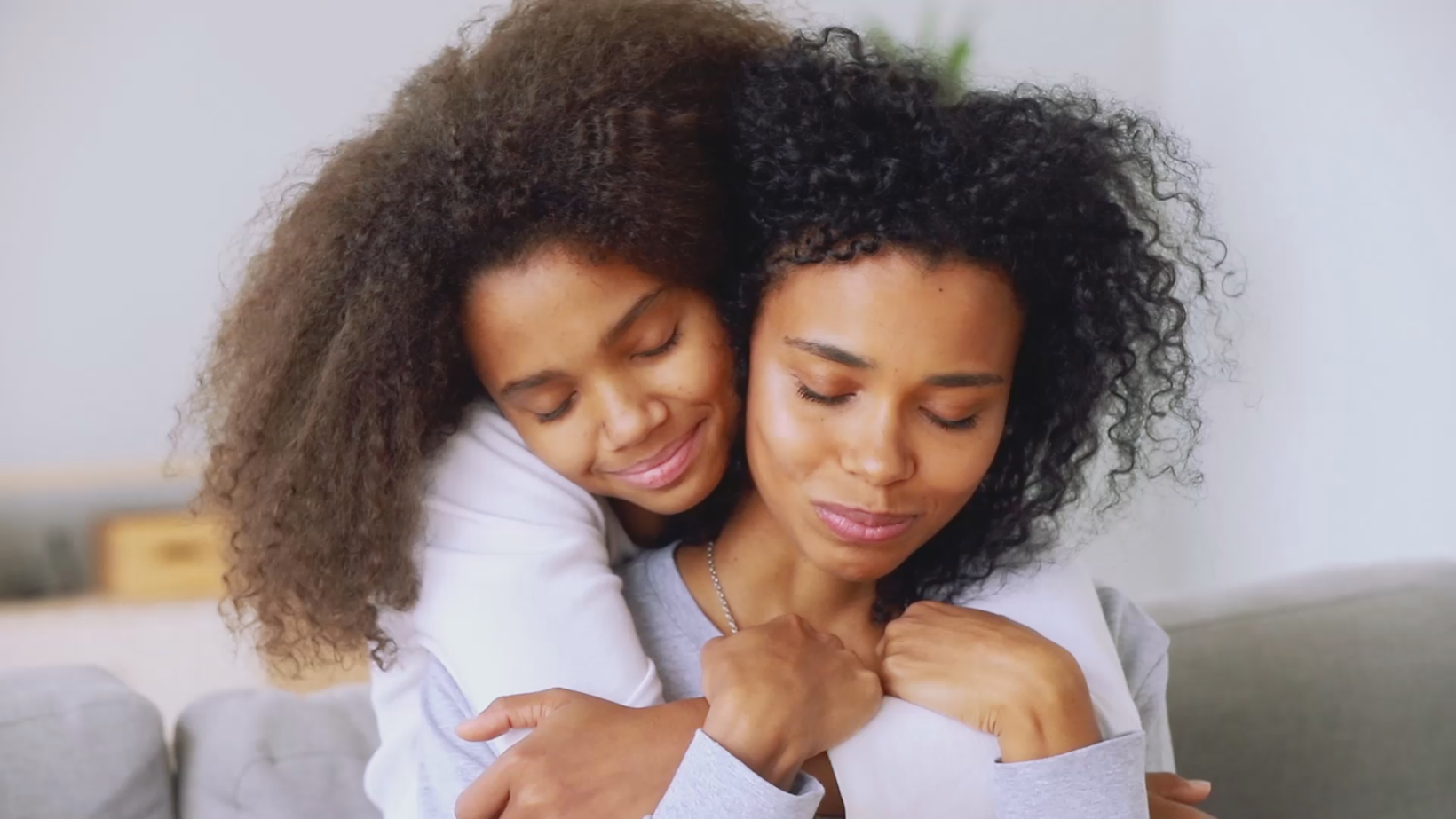 A touching scene of two women sharing an intimate moment. One woman with curly hair wraps her arms around the other, who has similar curly hair, as they embrace each other on a soft sofa. Both women have closed eyes and serene expressions, radiating warmth, comfort, and love. The soft lighting and neutral-toned surroundings enhance the sense of peace and affection between them.
