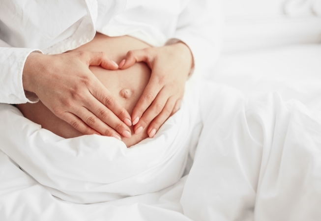 Close-up of a pregnant woman in white clothing making a heart shape with her hands over her bare belly while sitting on a bed.