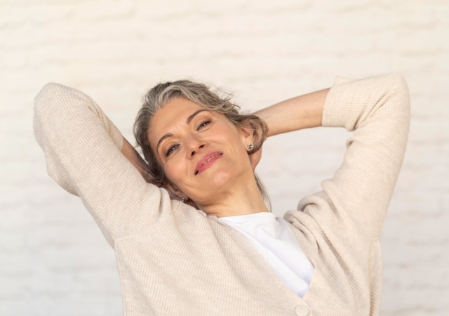 A smiling older woman stretching and relaxing, with her hands behind her head, wearing a light cardigan.