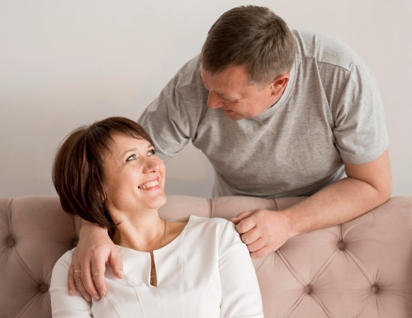 A smiling couple sitting together on a couch, with the man standing behind, gently resting his hand on the woman's shoulder.