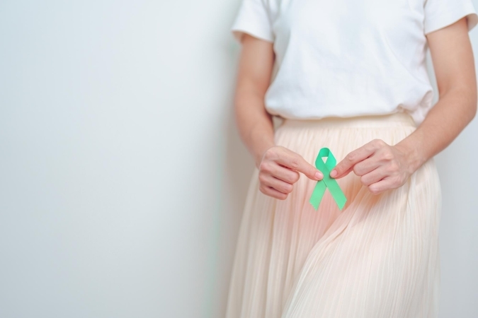 A woman wearing a white top and a light pleated skirt holds a teal awareness ribbon with both hands against a plain background.