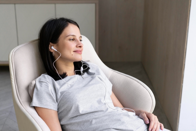 A woman in a gray t-shirt is sitting in a comfortable chair, listening to music with earphones and smiling calmly.